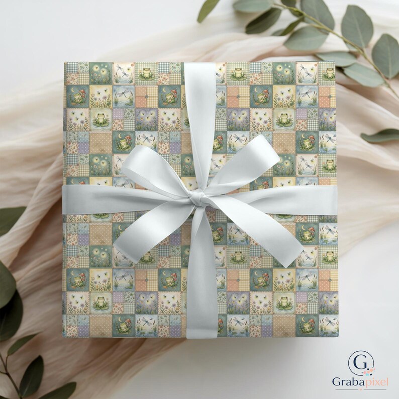 K&ouml;nnte beinhalten: Ein quadratisches Geschenk, verpackt in gemustertem Papier mit einem wei&szlig;en Satinband, das zu einer Schleife gebunden ist. Das Papier zeigt ein Patchwork-Design mit Illustrationen von Fr&ouml;schen, Libellen und Blumen in Pastellfarben. Die Schleife ist mittig auf dem Geschenk.