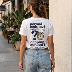 May include: A white short-sleeved t-shirt featuring dark blue text that reads normal bedtime? no, thanks. napping at noon? yes, please. The design includes a large question mark and an illustration of an analog alarm clock set to twelve.