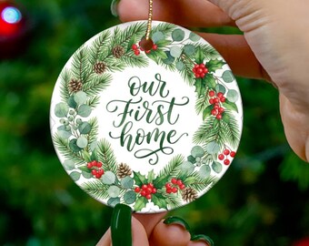May include: A round white ceramic ornament featuring a printed wreath of green pine needles, eucalyptus leaves, brown pinecones, and red berries. The center of the wreath displays the black cursive text "our first home" with a gold hanging cord.