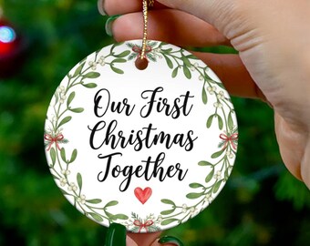 May include: A round white ceramic ornament featuring a green mistletoe wreath border and the black text "Our First Christmas Together" with a small red heart underneath. The ornament hangs from a gold string against a blurred holiday tree.