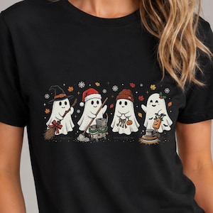 May include: A black short-sleeve t-shirt featuring a graphic print of four white ghosts. The ghosts are wearing various seasonal accessories including a witch hat, a Santa hat, a knit beanie, and a scarf, surrounded by small autumn leaves.