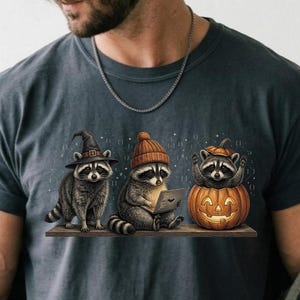 Op de afbeelding: Een antracietgrijs t-shirt met een afbeelding van drie wasberen. Eén wasbeer draagt een heksenhoed, de tweede draagt een muts en houdt een tablet vast, en de derde zit in een uitgesneden oranje pompoen.