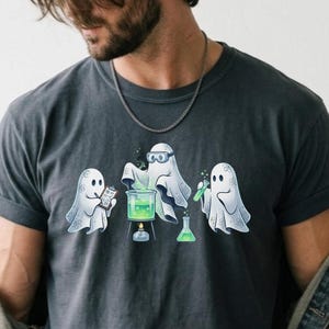 Peut inclure: Un t-shirt à col rond gris anthracite avec un motif représentant trois fantômes blancs réalisant une expérience scientifique. Les fantômes sont entourés d'équipement de laboratoire, dont un bécher au liquide vert fluorescent et un bec bunsen.