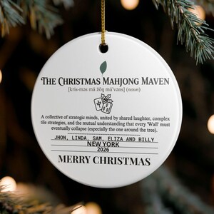 May include: A round white ceramic Christmas ornament hanging by a gold string. It features black text reading The Christmas Mahjong Maven, a definition, a mahjong tile graphic, names Jhon, Linda, Sam, Eliza and Billy, New York, 2026, and Merry Christmas.