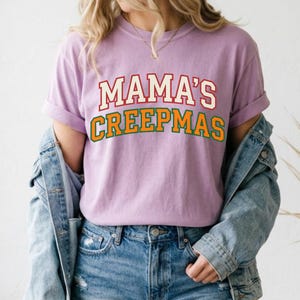 Op de afbeelding: Een lichtpaars T-shirt met korte mouwen met de tekst MAMA'S in het rood en CREEPMAS in het groen, beide in een vetgedrukt collegiaal lettertype. Het shirt wordt gedragen met een blauw spijkerjasje en een blauwe spijkerbroek voor een casual look.