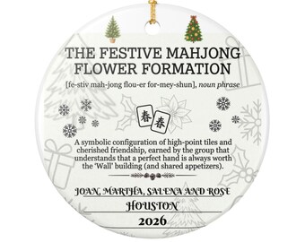 May include: A round white ceramic ornament with a gold hanging string. It features the text THE FESTIVE MAHJONG FLOWER FORMATION, a definition, two mahjong tiles, and holiday graphics including trees, snowflakes, and gifts. Names, city, and 2026 are printed.