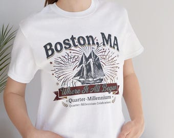 May include: White t-shirt featuring a graphic design with the text "Boston, MA" above a sailboat illustration. Below the sailboat is the text "Where It All Began" and "Quarter-Millennium Celebration".