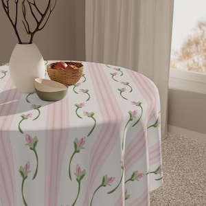 May include: A round tablecloth with a floral pattern and pink stripes. The tablecloth is on a round table with a white vase, two bowls, and a basket of apples. The floral pattern features pink buds and green stems.