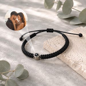 Op de afbeelding: Een zwarte gevlochten koordarmband met een centrale cilindrische zilverkleurige metalen kraal. De kraal bevat een kleine ronde projectielens die een foto van een knuffelend koppel toont. De armband heeft verstelbare uiteinden met trekkoorden.