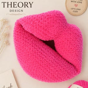Puede incluir: Un cojín de ganchillo de color rosa intenso con forma de labios. El cojín tiene una superficie texturizada y difusa. En la imagen se incluye una etiqueta redonda que dice "HANDMADE WITH LOVE LET'S STAY COZY" y un ovillo de lana con la etiqueta "THE YARN THEORY DESIGN".