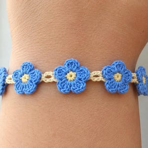May include: A crochet bracelet featuring a series of five blue flowers with small yellow centers, connected by a thin cream-colored yarn band. The floral design is crafted with visible yarn stitches, creating a textured, repeating pattern around the wrist.