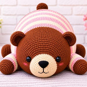 May include: A crochet bear plush toy featuring a dark brown head and limbs, a cream-colored muzzle, and a round body with horizontal stripes in shades of pink, cream, and white. The bear has black plastic eyes, a black nose, and pink cheek accents.