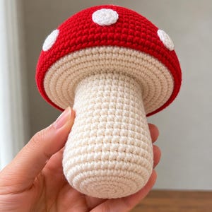 May include: A crocheted mushroom toy featuring a bright red cap with white circular spots and a cream-colored cylindrical stem. The texture is created with tight, uniform stitches throughout the entire structure, showcasing a rounded top and a flat base.