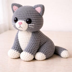 May include: A handmade crochet cat doll in gray and white. The cat has large black eyes, pink inner ears, and a pink nose. The cat is sitting with its tail extended and its paws are white.