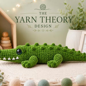 Könnte beinhalten: Ein grünes, gehäkeltes Krokodil-Stofftier mit strukturiertem Körper, einer Reihe kleiner Höcker auf dem Rücken, weißen Garnzähnen und großen dunklen Augen. Der Text THE YARN THEORY DESIGN ist über dem Spielzeug sowie ein Garnknäuel-Symbol zu sehen.