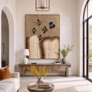 Puede incluir: Un gran cuadro rectangular que presenta un paisaje texturizado en beige y marrón con patrones abstractos de anillos de árboles y cinco tallos de flores delicados, cuatro en verde apagado y uno en azul vibrante, sobre un fondo dorado metálico.