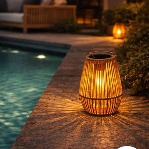 Puede incluir: Una linterna de bambú iluminada, con un brillo cálido, se encuentra sobre una superficie de piedra cerca de una piscina. La luz de la linterna proyecta un patrón de sombras. La escena está ambientada al aire libre al anochecer, con un fondo borroso.