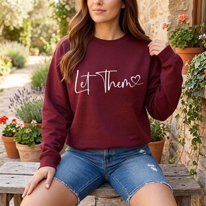 K&ouml;nnte beinhalten: Ein weinrotes Sweatshirt mit dem Schriftzug "Let Them" in wei&szlig;er Schrift und einem Herzsymbol. Das Sweatshirt wird mit Jeansshorts kombiniert. Das Bild ist im Freien mit Pflanzen und einer Steinmauer im Hintergrund aufgenommen.