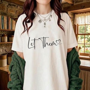 K&ouml;nnte beinhalten: Wei&szlig;es T-Shirt mit dem schwarzen Schriftzug "Let Them" mit einem Herz. Das Shirt wird mit einer gr&uuml;nen Strickjacke und mehrschichtigen Halsketten getragen.