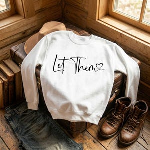 K&ouml;nnte beinhalten: Ein wei&szlig;es Sweatshirt mit dem Schriftzug "Let Them" in schwarzer Schrift, wobei ein Herz den Buchstaben "o" ersetzt. Das Sweatshirt wird auf einem Vintage-Koffer pr&auml;sentiert, mit braunen Stiefeln, einem Hut und Jeans im Hintergrund.