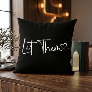 K&ouml;nnte beinhalten: Schwarzes quadratisches Kissen mit dem wei&szlig;en Text "Let Them" und einem Herzsymbol. Das Kissen liegt auf einer Holzoberfl&auml;che mit einer Pflanze und B&uuml;chern im Hintergrund. Dekoratives Wohnaccessoire.