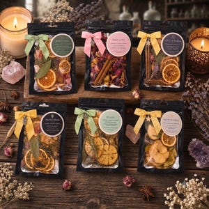 May include: Six black resealable pouches containing dried botanical blends, including orange slices, cinnamon sticks, and flower petals. Each pouch features a colored ribbon bow with a small metal charm and a circular label with text.