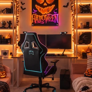 Puede incluir: Una silla de juego negra con detalles de luz LED azul y morada colocada frente a un escritorio con dos monitores. Sobre el escritorio, un letrero de pared rectangular presenta una cara de calabaza naranja brillante y la palabra HALLOWEEN en neón.