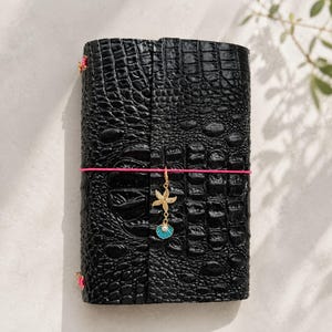 以下が含まれることがあります： 黒い革のジャーナル。クロコダイル柄のテクスチャ。ピンク色のゴムバンドがジャーナルを包んでいます。ヒトデ、真珠、ターコイズの貝殻が付いたゴールドチャームがバンドからぶら下がっています。ピンクの花のアクセントが背表紙にあります。