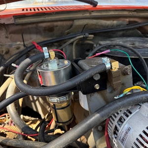May include: A cylindrical fuel filter with a clear bottom housing and a metal top, mounted on a black bracket within an engine bay. The unit features black rubber hoses secured with metal clamps, brass fittings, and various electrical wires.
