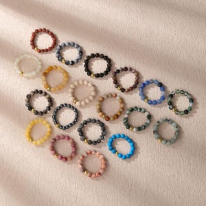 May include: A collection of beaded bracelets in various colors, including red, black, yellow, and blue. Each bracelet features small, round beads and gold-colored accents. The bracelets are arranged on a light-colored surface.