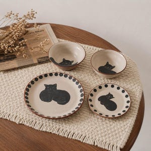 May include: A set of two ceramic bowls and two ceramic plates in a cream color, each featuring a black cat illustration. The plates have a black polka dot border. The bowls show a black cat design on the interior. The items are arranged together.