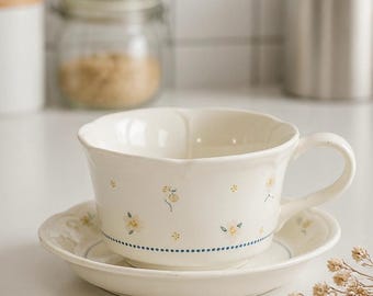 Juego de taza y platillo de cerámica con diseño floral pintado a mano, color marfil con borde festoneado.