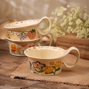 May include: Three off-white ceramic bowls with large handles, stacked on a burlap surface. The bowls are decorated with colorful floral and fruit designs. The bowls are part of a kitchenware set.