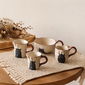 May include: A set of three off-white ceramic mugs and one matching bowl, each featuring a hand-painted black cat illustration. The pieces have irregular, wavy rims and dark brown handles, showcasing a rustic, handcrafted aesthetic.