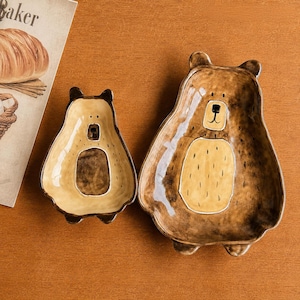 May include: Two bear-shaped ceramic serving dishes. The larger dish is brown with a yellow belly and a bear face. The smaller dish is avocado-shaped with a bear face. Both dishes are on a brown surface.