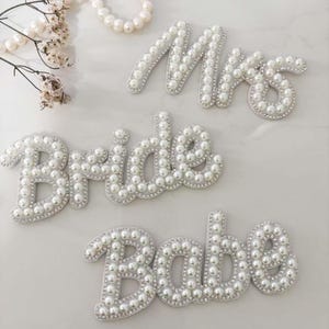 May include: Decorative pearl-covered word cutouts spelling "Mrs", "Bride", and "Babe". The letters are outlined with small, clear rhinestones. A string of pearls and floral accents are in the background. Ideal for wedding decor or bridal showers.