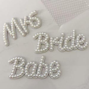 May include: White pearl-covered letters spelling "Mrs", "Bride", and "Babe". The letters are outlined with small, clear rhinestones. The letters are likely for a bridal shower or wedding decoration.