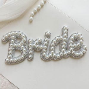 Può includere: Un'opera d'arte decorativa con la scritta "Bride" bianca, impreziosita da perle finte e strass. Le lettere sono bordate d'argento e poste su uno sfondo bianco, con una fila di perle nell'angolo in alto a destra. Tema nuziale.
