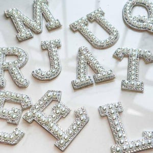 Puede incluir: Variedad de letras adornadas con perlas, incluyendo M, U, R, J, A, T, V y L. Cada letra está bordeada con pequeños diamantes de imitación transparentes. Las perlas son blancas y las letras están sobre una superficie rosa claro.