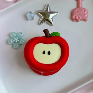 May include: A red apple-shaped phone grip with a cream-colored center, a brown stem, and a green leaf. Surrounding the apple are a silver star, a light blue flower-shaped charm, and a pink cat-shaped charm on a white surface.