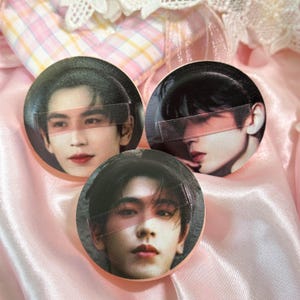 May include: Three circular cosmetic powder puffs, each featuring a printed portrait of a person with dark hair. A semi-transparent horizontal band crosses the eyes of each individual in the portraits. The puffs have a soft, rounded shape.