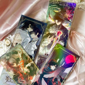 May include: Four rectangular anime-style art prints featuring characters with dark hair in elaborate outfits. One print shows a figure in a witch hat, another depicts two figures in ruffled collars, and others show characters with wings and red accents.