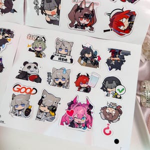 May include: A sticker sheet featuring various chibi-style anime characters in dynamic poses and expressions. The designs include a panda eating, a character holding a sign with Chinese text, a pink-haired figure with a heart, and the word GOOD.