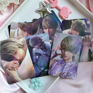 May include: A collection of rectangular trading cards featuring anime-style illustrations of a character with light hair. The cards display various poses and outfits, including a purple ensemble with star accents, and are arranged on a white tray.