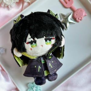 May include: A plush doll featuring black hair with lime green highlights, pale skin, and embroidered green eyes. The doll wears a dark purple jacket with lime green accents and small pocket details. The character has a neutral expression with small red markings under the eyes.