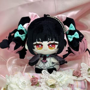 May include: A plush doll featuring black hair styled in twin tails with light pink and mint green bows. The doll has red eyes, a pale face, and wears a white and black outfit with a small cape. A silver ball chain is attached to the top of the head.