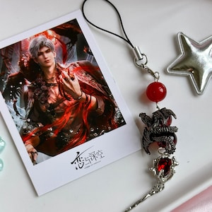 May include: A rectangular photo card featuring a male character with silver hair in a red and black outfit, with the text "恋与深空" at the bottom. Attached is a black cord charm with a red bead, a black dragon charm, and a silver-toned pendant with a red gem.