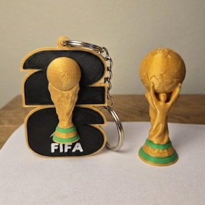 May include: A black and gold keychain featuring the number 23 with a gold FIFA trophy emblem and a separate small gold and green FIFA trophy figurine. The keychain has a metal ring and chain attached to the top of the black number 23 base.