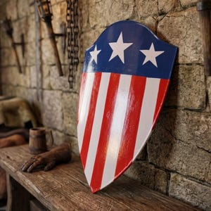 May include: A heater-style shield featuring a glossy finish with a blue upper section adorned with three white five-pointed stars, and a lower section displaying vertical red and white stripes. The shield has a pointed base and curved top edges.