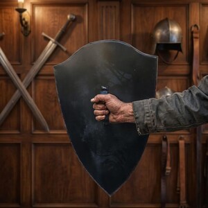 May include: A heater-style shield with a smooth, matte black finish and a central vertical handle. The shield features a pointed base and a curved top edge, presenting a solid, dark surface that contrasts with the metallic sheen of the handle.